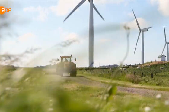 Ausschnitt eines ZDF-Beitrags, auf dem man blauen Himmel über einer Wiese mit Windrädern und einem Trecker sieht.