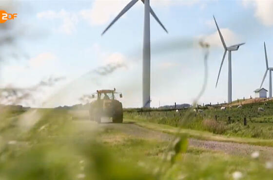 Ausschnitt eines ZDF-Beitrags, auf dem man blauen Himmel über einer Wiese mit Windrädern und einem Trecker sieht.