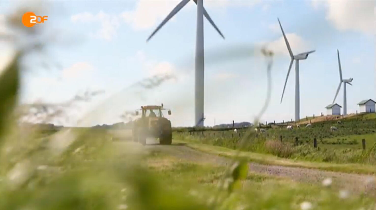 Ausschnitt eines ZDF-Beitrags, auf dem man blauen Himmel über einer Wiese mit Windrädern und einem Trecker sieht.