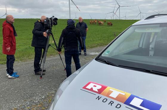 Kameraleute filmen vor einem grünen Feld mit Kühen und Windrädern im Hintergrund den Geschäftsführer des Dirkshofs, Dirk Ketelsen. Im Vordergrund ist ein RTL-Auto zu sehen.