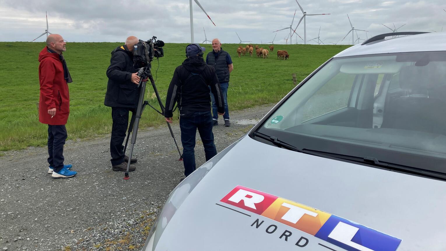 Kameraleute filmen vor einem grünen Feld mit Kühen und Windrädern im Hintergrund den Geschäftsführer des Dirkshofs, Dirk Ketelsen. Im Vordergrund ist ein RTL-Auto zu sehen.