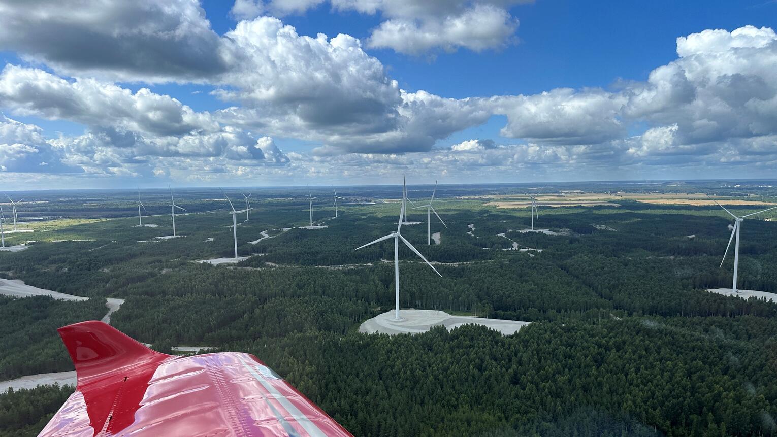 Aus dem Fenster eines kleinen Passagierflugzeugs fotografiert, von dem man den roten Flügel sieht, sieht man dichte grüne Wälder, dazwischen Flüsse und Windräder.