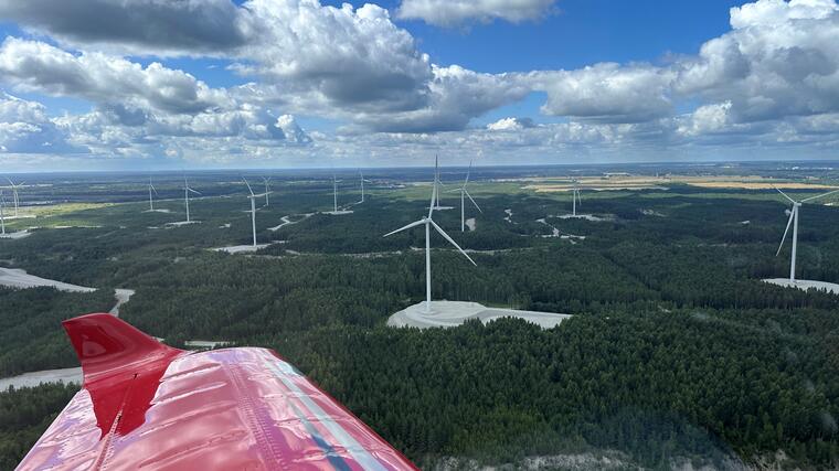 Aus dem Fenster eines kleinen Passagierflugzeugs fotografiert, von dem man den roten Flügel sieht, sieht man dichte grüne Wälder, dazwischen Flüsse und Windräder.
