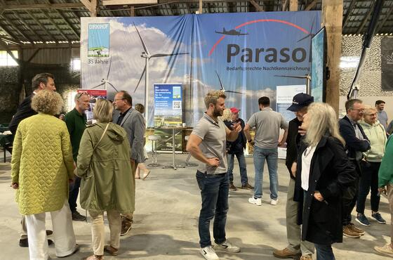 Menschen stehen beim Sönke-Nissen-Koog-Hoftag in Grüppchen verteilt in einer Halle und sprechen miteinander. Im Hintergrund ein großes Plakat mit der Aufschrift PARASOL.