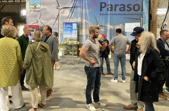 Menschen stehen beim Sönke-Nissen-Koog-Hoftag in Grüppchen verteilt in einer Halle und sprechen miteinander. Im Hintergrund ein großes Plakat mit der Aufschrift PARASOL.