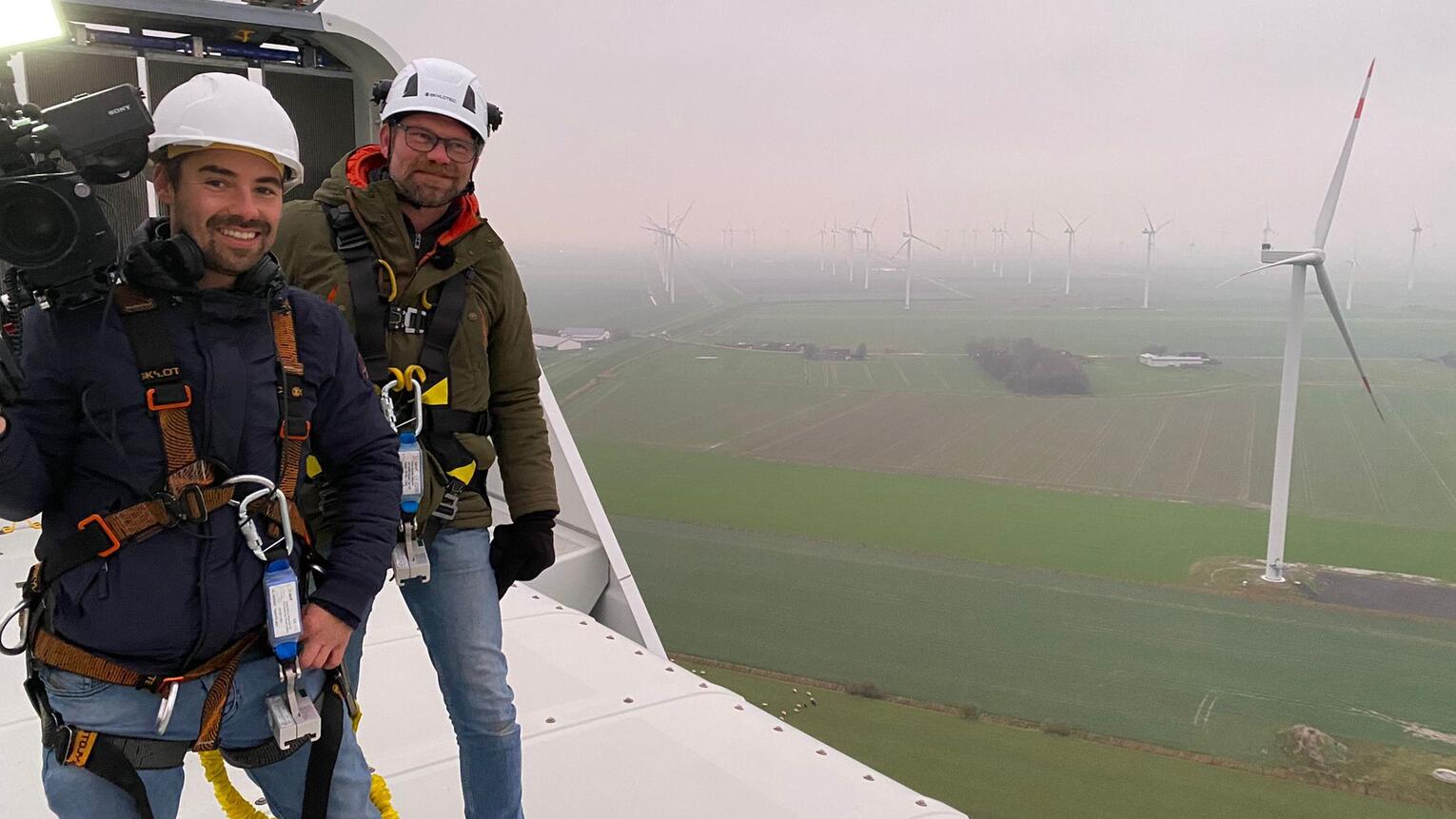 Zwei Männer mit Kameraausrüstung stehen bei nebligem Wetter auf einer Windturbine und lächeln.