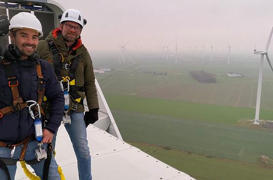Zwei Männer mit Kameraausrüstung stehen bei nebligem Wetter auf einer Windturbine und lächeln.