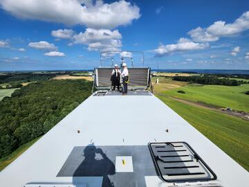 Bei strahlend blauem Himmel stehen zwei Personen auf einer Windturbine, im Hintergrund grüne Wiesen und Wälder.