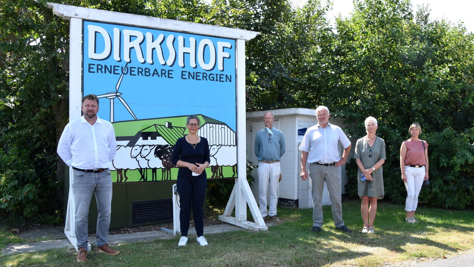 Nina Scheer, eine Frau mittleren Alters mit Brille, dunklem Haar und dunklem Longsleeveshirt, steht mit mehreren anderen Personen vor einem großen Schild mit dem Logo des Dirkshofs am Eingang des Bürgerwindparks. Im Hintergrund sind Büsche und Bäume.