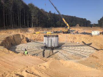Eine sandige Baustelle in einem Wald mit dem Fundament eines Windrads und Arbeitsgeräten ringsum.