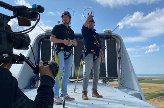 Ein Kameramann filmt, wie zwei Männer mit Schutzhelmen auf einer Windturbine stehen und sich umsehen. Einer der beiden Männer zeigt auf etwas.