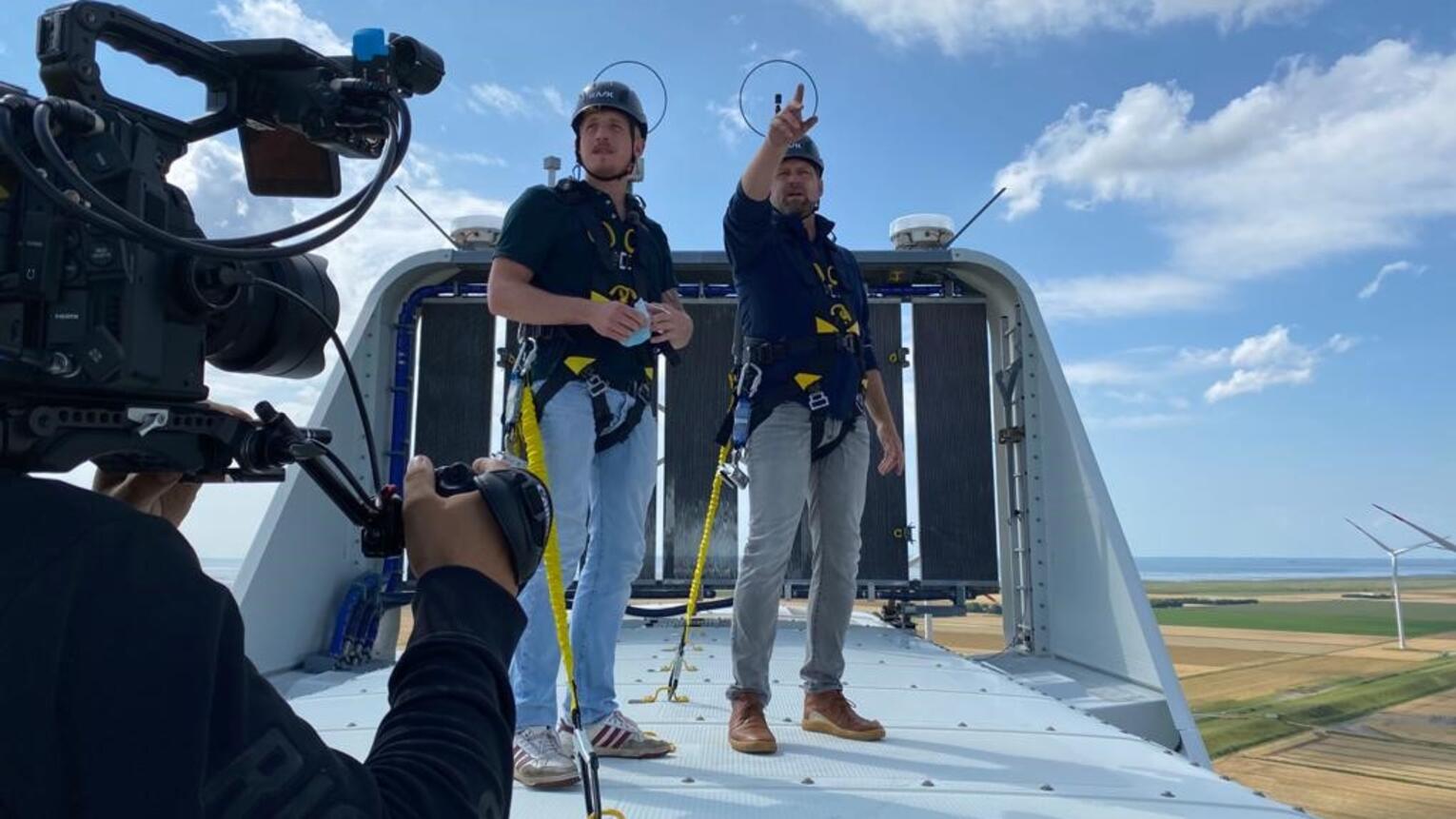 Ein Kameramann filmt, wie zwei Männer mit Schutzhelmen auf einer Windturbine stehen und sich umsehen. Einer der beiden Männer zeigt auf etwas.