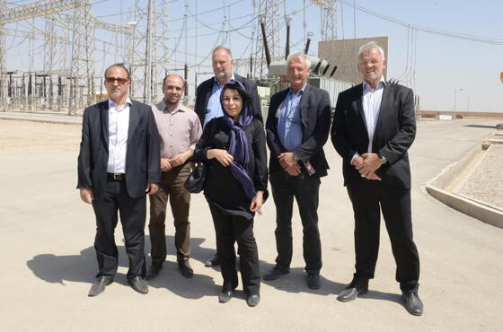 Fünf Männer und eine Frau stehen an einem sonnigen Tag in Businesskleidung vor einem Kraftwerk und posieren für ein Gruppenfoto.
