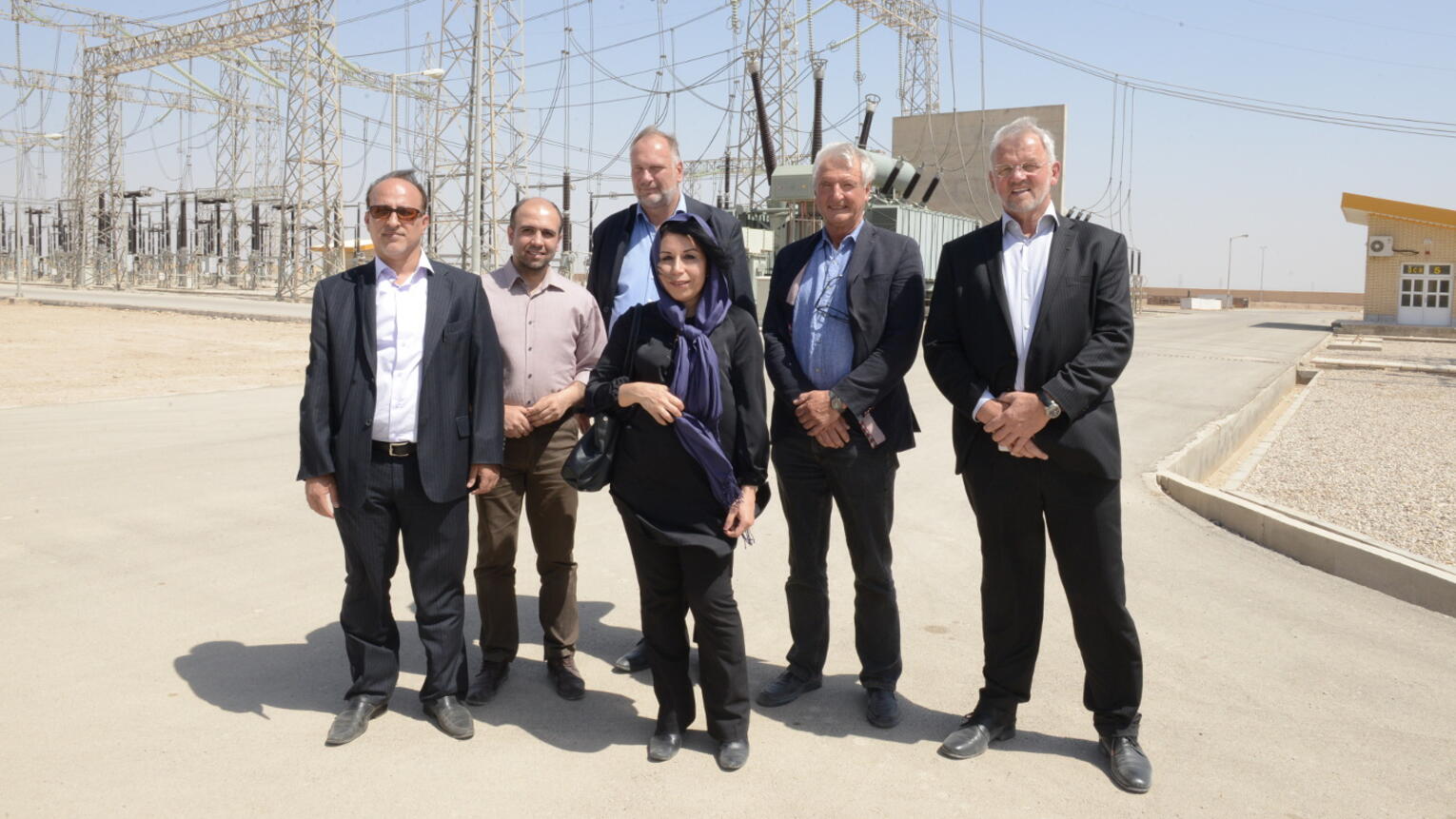 Fünf Männer und eine Frau stehen an einem sonnigen Tag in Businesskleidung vor einem Kraftwerk und posieren für ein Gruppenfoto.