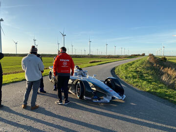 Ein Rennwagen auf einer Landstraße, daneben Männer, die ihn sich ansehen. Im Hintergrund Felder und Windräder.