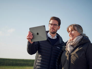 Ein Mann und eine Frau, die sich draußen auf einem Feld auf dem Tablet ein Video angucken.