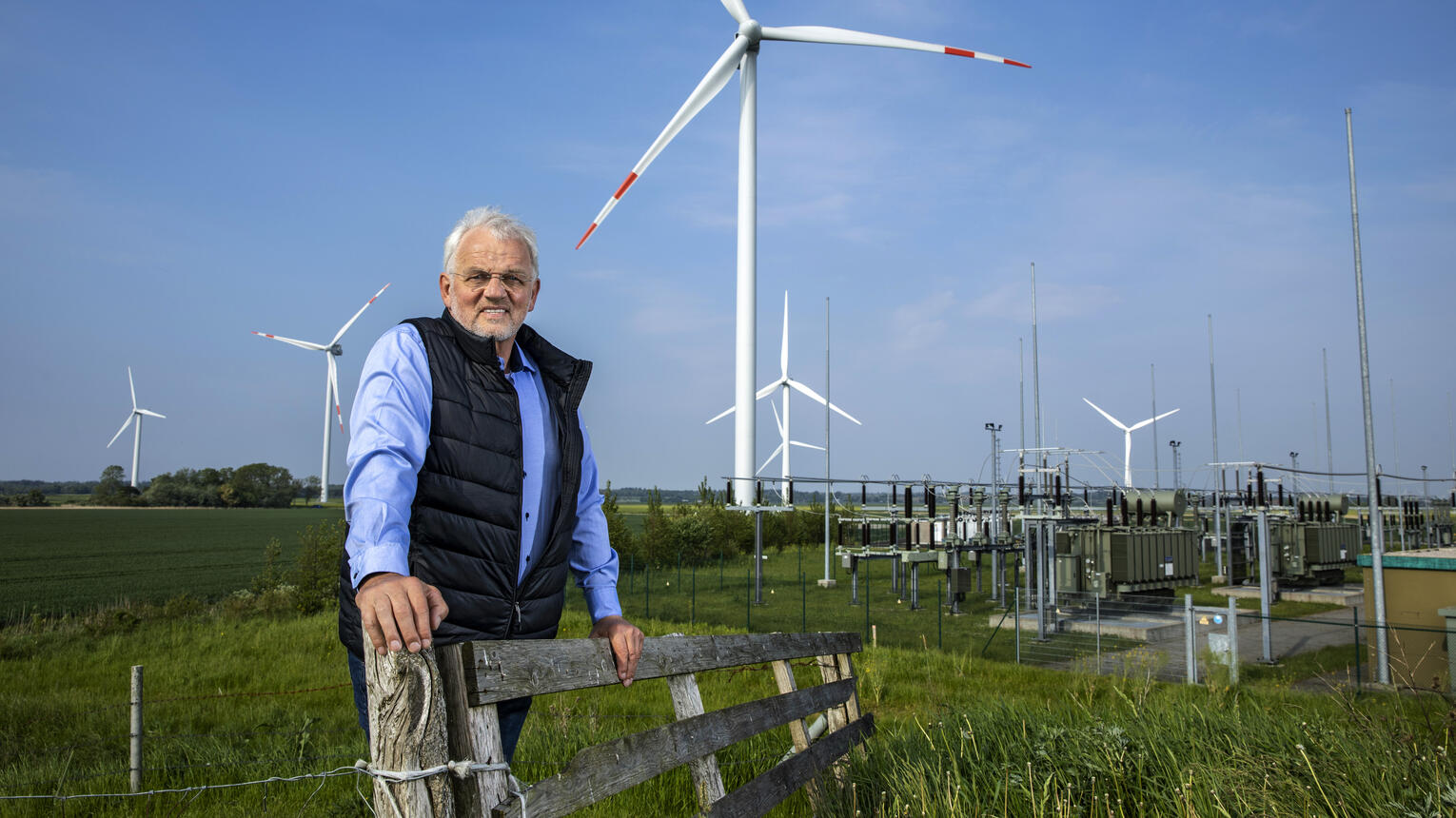 Geschäftsführer Dirk Ketelsen lehnt im Windpark an einem Zaun. Im Hintergrund blauer Himmel, Windräder und grüne Wiesen.