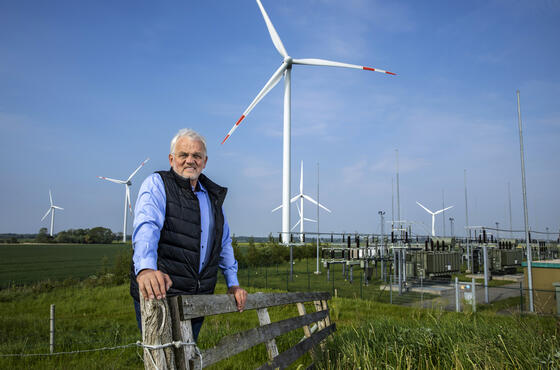 Geschäftsführer Dirk Ketelsen lehnt im Windpark an einem Zaun. Im Hintergrund blauer Himmel, Windräder und grüne Wiesen.
