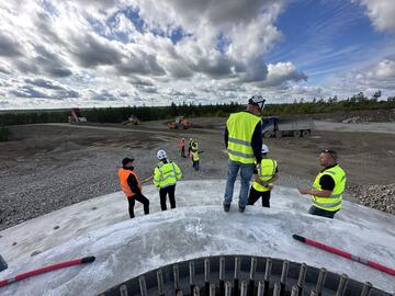 Auf der Baustelle eines Windrads stehen mehrere Bauarbeiter in leuchtenden Schutzwesten und unterhalten sich.