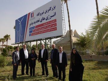 Mehrere Personen posieren vor einem arabisch beschrifteten Schild inmitten von Palmen für ein Gruppenfoto.