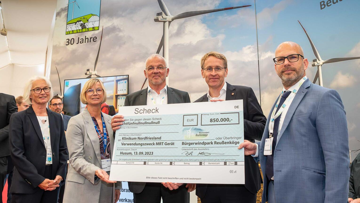 Vier Menschen, darunter Dirk Ketelsen und der Ministerpräsident, halten einen großen Scheck mit der Summe 850.000 Euro in die Kamera. Thoma Ketelsen steht links daneben. Im Hintergrund ein großes Plakat mit Windrädern darauf.