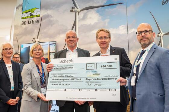 Vier Menschen, darunter Dirk Ketelsen und der Ministerpräsident, halten einen großen Scheck mit der Summe 850.000 Euro in die Kamera. Thoma Ketelsen steht links daneben. Im Hintergrund ein großes Plakat mit Windrädern darauf.