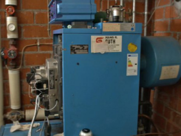 Screenshot eines Sendungsausschnitts, in dem eine blaue Maschine vor einer Backsteinwand zu sehen ist.