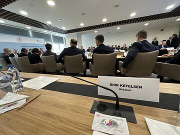 Ein Konferenzraum voller Menschen, mit Tischen und Bürostühlen, fotografiert von einem der Plätze aus. Am Platz steht ein Namensschild und ein paar Notizzettel liegen bereit.