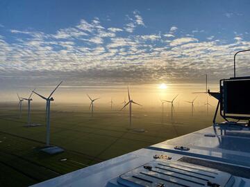 Blick von einer Windturbine aus auf die darunter liegende Landschaft. Nebel, lockere Wolken und strahlender Sonnenschein hüllen die grünen Felder und anderen Windräder in der Ferne in ein warmes Leuchten.