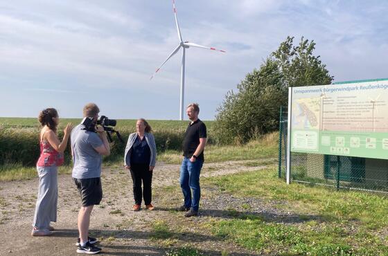 Vier Personen stehen sich am Eingang des Bürgerwindparks Reußenköge paarweise gegenüber, ein Mann filmt die anderen.