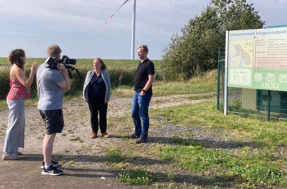 Vier Personen stehen sich am Eingang des Bürgerwindparks Reußenköge paarweise gegenüber, ein Mann filmt die anderen.