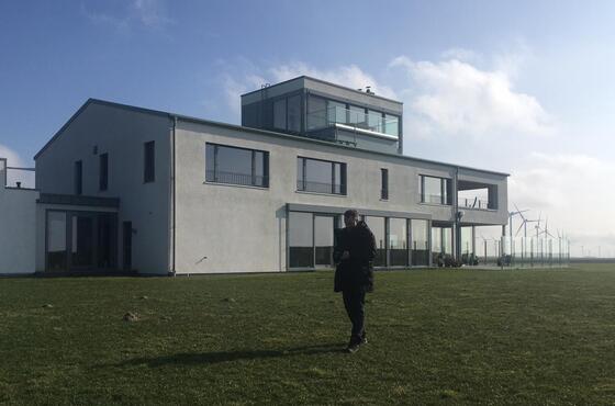 Eine Person geht bei Sonnenschein auf einer weitläufigen Grünfläche vor einem modernen Gebäude vorbei.