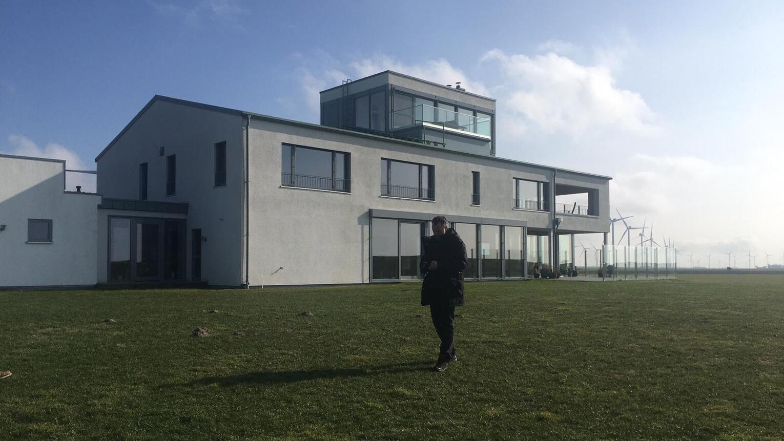 Eine Person geht bei Sonnenschein auf einer weitläufigen Grünfläche vor einem modernen Gebäude vorbei.