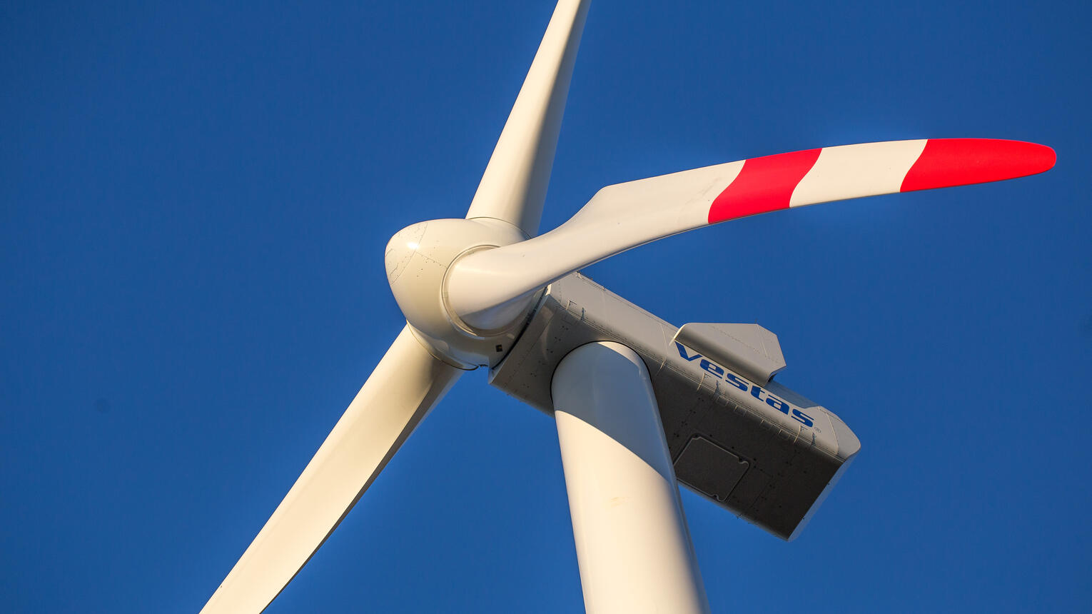 Ein weißes Windrad des Herstellers VESTAS mit roten Streifen an den Rotorblättern in Nahaufnahme vor einem blauen Himmel.
