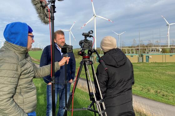 Ein Kameramann mit blauer Kapuze filmt ein Interview zwischen einem Mann und einer Frau in einem Feld.