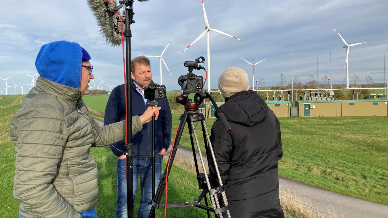 Ein Kameramann mit blauer Kapuze filmt ein Interview zwischen einem Mann und einer Frau in einem Feld.