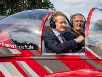 Der Ministerpräsident und ein Pilot im Cockpit einer kleinen, roten Passagiermaschine, das Verdeck gerade offen.