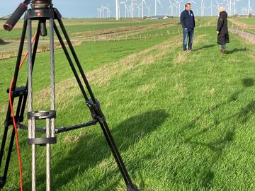 Ein Mann wird auf einem Feld von einer Frau interviewt, im Vordergrund steht ein Tripod mit einer Kamera und filmt sie.
