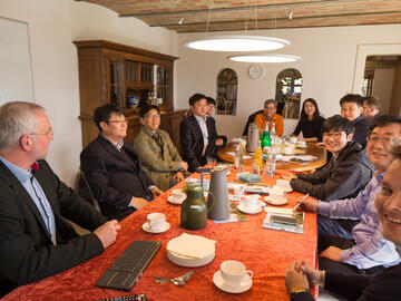 Koreanische Besucher sitzen in einem Innenraum des Dirkshofs mit den Gastgebern an einem langen Tisch, lachen und nehmen Getränke zu sich.