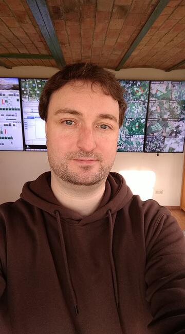 Ein Mann vor einer Wand mit Plänen lächelt in die Kamera. Es ist ein Selfie.