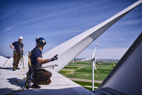 Zwei Arbeiter mit Schutzhelmen, gesichert durch Seile, arbeiten auf der Turbine eines Windrads und sprechen etwas ab.