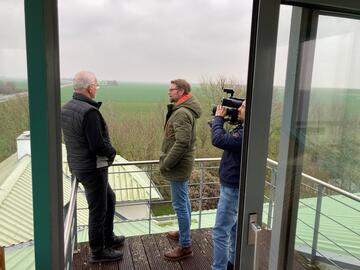 Drei Männer stehen bei nebligem Wetter auf einem Balkon, einer filmt, zwei andere sprechen miteinander. Im Hintergrund grüne Wiesen.
