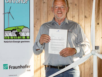 Mann steht mit Zertifikat vor einer Holzwand und lacht, daneben ein Aufsteller mit der Aufschrift „Dirkshof – Parasol“ sowie Logos offizieller Förderungen, im Vordergrund ein Miniatur-Windrad.