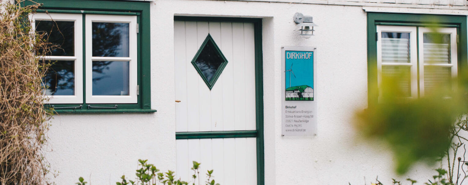 Eine weiße Tür mit einem kleinen Fenster an einem weißen Bauernhaus, im Vordergrund grüne Büsche. Neben Tür hängt ein Schild, auf dem „Dirkshof“ steht.