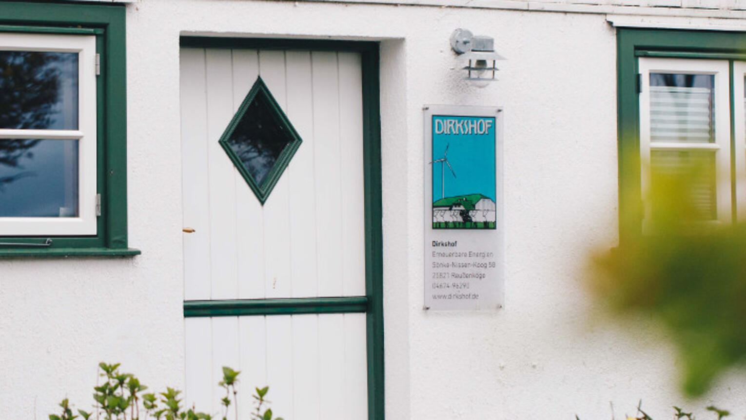 Eine weiße Tür mit einem kleinen Fenster an einem weißen Bauernhaus, im Vordergrund grüne Büsche. Neben Tür hängt ein Schild, auf dem „Dirkshof“ steht.