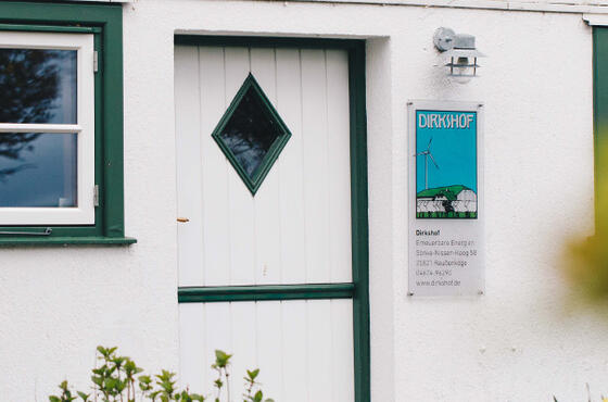 Eine weiße Tür mit einem kleinen Fenster an einem weißen Bauernhaus, im Vordergrund grüne Büsche. Neben Tür hängt ein Schild, auf dem „Dirkshof“ steht.