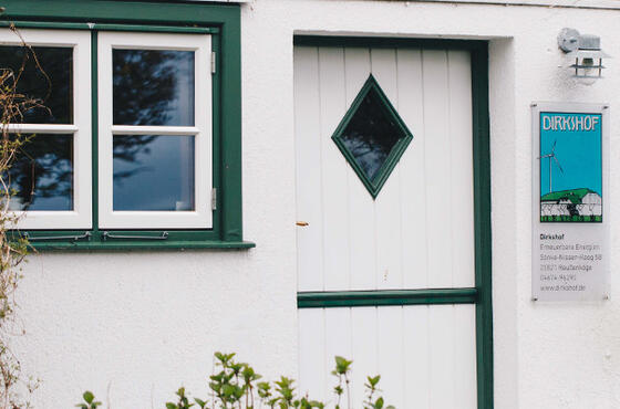 Eine weiße Tür mit einem kleinen Fenster an einem weißen Bauernhaus, im Vordergrund grüne Büsche. Neben Tür hängt ein Schild, auf dem „Dirkshof“ steht.