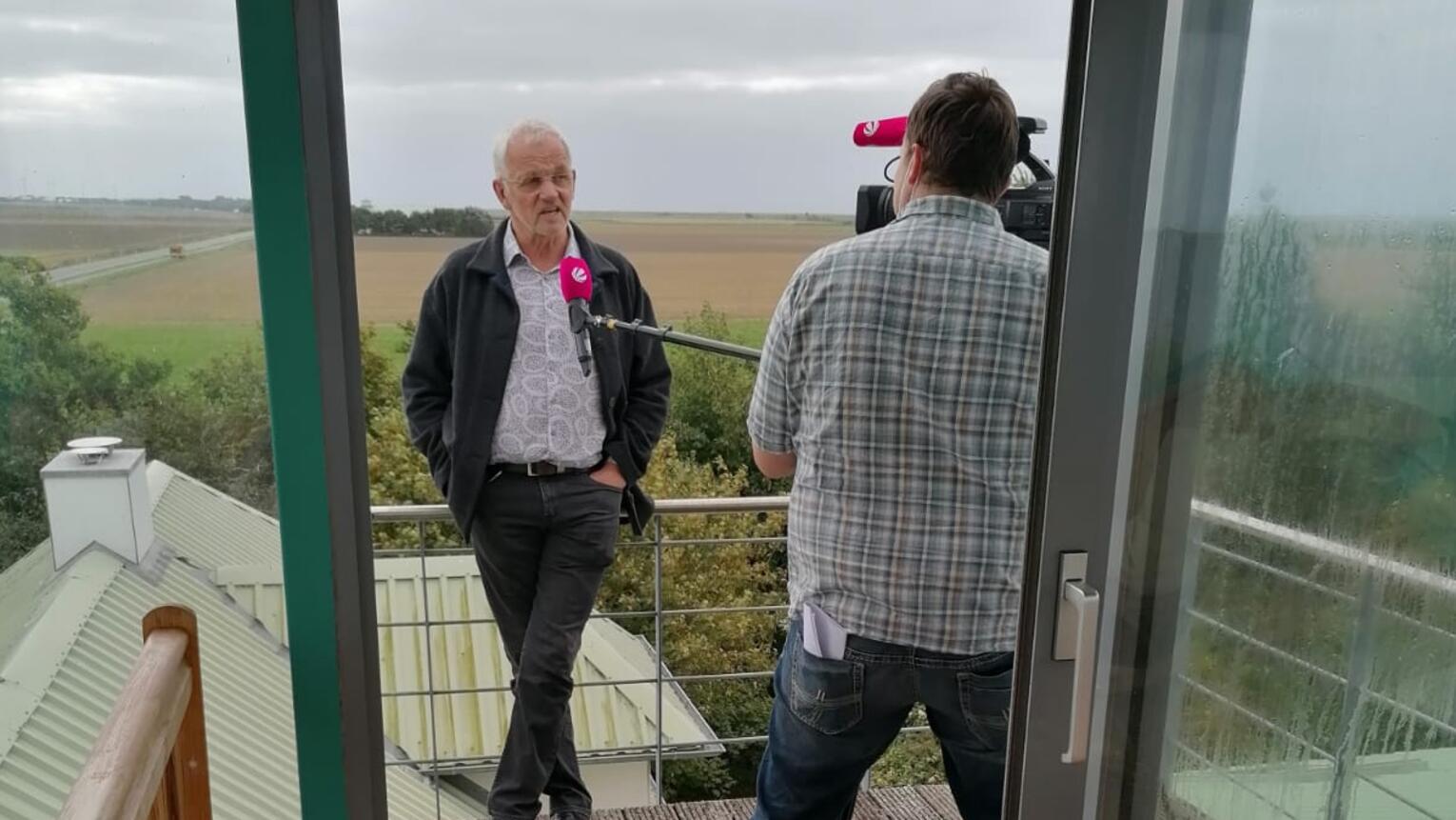 Blick aus einem Innenraum auf einen Balkon, auf dem ein älterer Mann vor der Kamera steht und interviewt wird.
