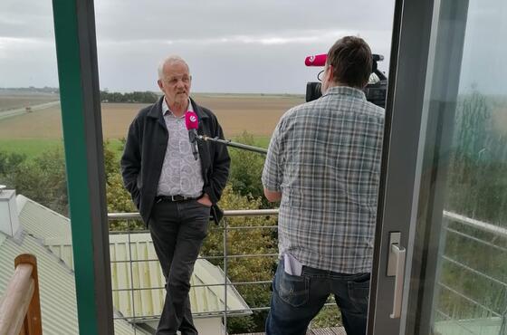 Blick aus einem Innenraum auf einen Balkon, auf dem ein älterer Mann vor der Kamera steht und interviewt wird.