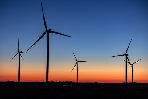 Dunkle Silhouetten von Windrädern vor einem orange-blauen Abendhimmel. An den Turbinen der Windräder blinkt es rot.
